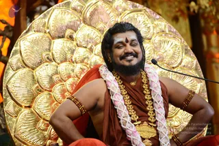 A smiling man seated in front of a large golden lotus-petal backdrop, with a microphone on a stand at the right. He wears a rust-orange shawl over his body, a double strand of pink and white flower garland, layered gold necklaces, beaded bracelets on his upper arms, and has a red bindi and white markings on his forehead. His hands rest on his hips and he looks sideways with a broad grin. Warm orange lighting fills the background.