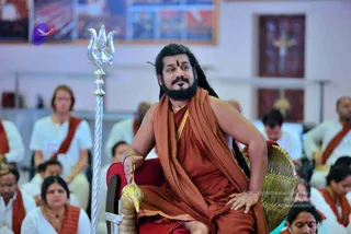 A man with long dark hair and a full beard sits on an ornate gold-and-red throne-style chair in an indoor hall, wearing a saffron-orange shawl wrapped over one shoulder, a gold necklace, and a red-brown dhoti-style garment, with a vermilion mark on his forehead. He rests one hand on the armrest and holds a tall silver trident beside him. Seated audience members in white clothes with orange-red shawls fill the foreground and background, and a framed picture hangs on the wall behind. The seated man in saffron with long hair and a trident matches the page's named teacher, so this appearance-based label is a person, seated in a teaching role before the audience.