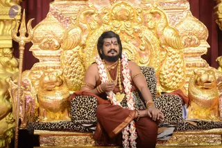 A bearded man with long swept-back dark hair sits on an ornate gold throne carved with lions and scrollwork, flanked by golden lion figures and a tall gold trident on the left. He wears a rust-brown robe, a gold chain with a pendant, gold armlets and bangles, and a white and red flower garland. He holds a small object in one hand while resting the other on the leopard-print cushion. Swamiji (probable) cues here are the long hair, ornate gold necklaces and saffron-toned robe, with a seated ceremonial pose on the throne; this is an in-person candidate on the dais.