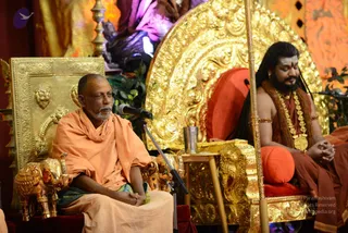 Two men sit on ornate gold thrones in a temple-like hall with a gold-framed backdrop and red-orange cushions. On the left, an older bald man with a grey beard wears an orange-saffron shawl and a green-striped cloth, speaking into a microphone on a stand with his hands clasped. On the right, a man with long dark hair, a dark beard, and white tilak marks sits in meditative posture, wearing a saffron-orange robe and multiple gold necklaces, with hands folded. A metal tumbler sits on a small table between them, and a long golden staff stands beside the right throne. Gold elephant figures decorate the arms of the left throne. A purple-draped deity figure is visible in the background.