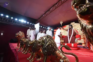 A photograph of a stage event under a red canopy with stage lights overhead. A seated man with long dark hair and a beard wears a rust-orange robe and a white flower garland, sitting on an ornate gold throne with red cushions. A young man in white clothing holds up a silver trident beside him, and another person in orange stands at the right edge. Several white-clad young men stand in a line at the back left. In the foreground, a row of large gold-toned lion statues lines the red carpeted platform.