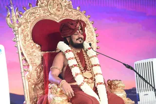 A photograph of a seated man on an ornate gold-and-red throne with a trident-topped finial, speaking into a microphone on a stand at right. He wears a maroon turban with a red tilak on the forehead, a dark-red saffron-style robe, layered gold necklaces, a large white and red flower garland, and gold bangles. Behind him is a pink-to-purple sky backdrop with a small golden deity murti, and a white air conditioning unit sits at the right edge.