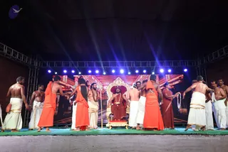A night stage event under a dark tent roof with blue and white stage lights. A seated figure in a red-brown robe and rudraksha-style beads sits on an ornate gilded throne with red cushions, set against a backdrop of a temple-like palace hall. Around him, devotees in white dhotis with gold borders, some bare-chested, and others in saffron-orange shawls and robes stand on a green-carpeted stage, many with their backs to the camera, bending or reaching toward the throne. A woman in a red-brown sari stands at right holding a cloth.