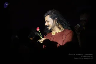 A photograph taken in a dark setting, with a single figure lit by a spotlight from the side. A man with long, swept-back dark hair and a full beard wears a rust-brown kurta with a gold pendant necklace. He looks down and smiles while holding a single pink rose with green stem and leaves in his hands. Behind him, out of focus, a second person stands at the right edge holding a small white flower, with a forehead marking visible. The background is almost entirely black.