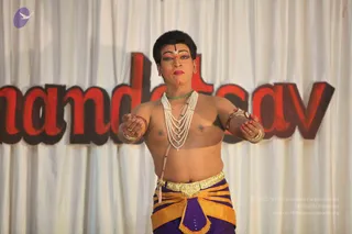 A photograph of a male dancer performing on stage in front of a white draped curtain with large red and brown letters reading "nanda...sav" behind him. He is bare-chested, wearing layered pearl necklaces, a green beaded collar, gold armbands and bangles, and a gold-buckled belt over a purple and orange patterned dhoti. His face has bold stage makeup with red lips, dramatic eyeliner and a bindi, and he holds his hands out in a dance gesture. Visible evidence shows a single performer; no candidate is identified from appearance, so no a person candidate is claimed.