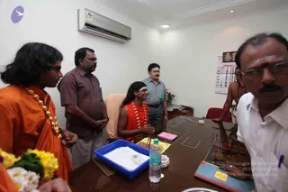 An indoor office photograph with a wall-mounted air conditioner and a January calendar on the wall. A man with long dark hair, wearing an orange robe and a gold-bead necklace, stands at left with flowers. Seated at a wooden table is a person in an orange robe with a gold-bead garland and a white tilak on the forehead, smiling with palms pressed together. Around them stand men in ordinary shirts and trousers, one in a grey shirt and one in a brown shirt. A man in white shirt and glasses sits at the right edge, and a bare-chested man with rudraksha beads appears at the back. Blue tray with papers and a water bottle sit on the table.