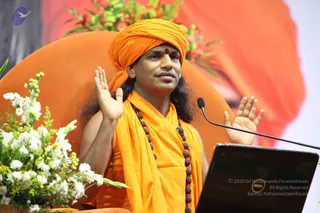 A man seated on an orange-upholstered chair before a microphone stand and a dark laptop, with a bouquet of white and yellow flowers at the left. He wears an orange turban with a red bindi, an orange shawl over orange clothing, and long brown rudraksha malas. He raises both palms in an open gesture while speaking. An orange and red backdrop fills the background.