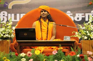 A seated man in saffron-orange robes and an orange turban sits on a large orange throne-style chair during a satsang discourse. He wears a long rudraksha mala and a red bindi, and a microphone stands before him. A dark laptop and a small speaker sit on a wooden table in front of him. Vases of white, pink and yellow flowers and orange gerbera flowers frame the stage, and a banner with Tamil lettering stands behind him.