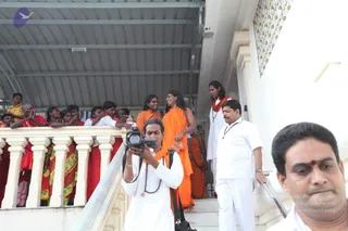 A photograph of a white stairway and balustrade beneath a metal roof. A man in white clothes with a rudraksha mala and red tilak holds a video camera to his eye, facing the stairs. Behind him, a figure in orange robes with long dark hair stands on the steps, and beside him another person in orange with a shawl stands with his head turned, smiling. A man in white shirt and trousers stands to the right on the steps, and a close-up man with a red bindi fills the lower right corner. Several people in red, yellow and orange garments stand along the upper balcony railing, many watching the scene, with a white tiled wall at the upper right.