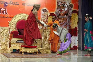 A figure in a red draped garment and tan head wrap stands beside a large ornate golden throne, reaching out to take papers from a saffron-robed man. A woman in a grey shawl over a purple skirt with green border bends forward holding papers, while a woman in a teal and pink sari with white flower adornments stands at right. Behind them a large orange backdrop shows a golden metallic deity murti with a crown, jewelled necklaces and garlands of yellow and red flowers, with Kannada script at the top.