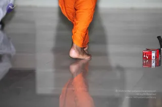 A close photograph of a bare foot and lower leg in loose orange cloth trousers, stepping across a polished grey floor that reflects the foot and cloth. Only the leg and foot of one person are visible at the top of the frame. On the right edge sits a red Sony VHS-style video cassette labelled 60, with a second cassette behind it. A faint white-clad figure appears at the far left edge. No face is visible, so no one can be identified from appearance.
