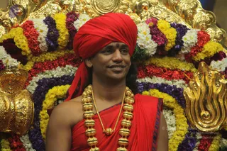 A smiling man in a red turban and red robe stands before a flower-decorated golden throne backdrop. He wears a heavy gold bead necklace of large ornaments over his bare chest and a gold chain, with a red tilak mark on his forehead. Behind him are garlands of yellow, white, red and purple flowers, and gold ornamental finials frame the throne.