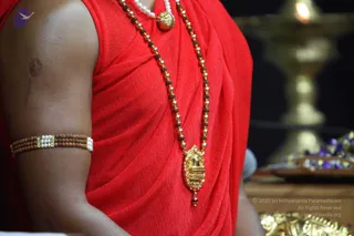 Close photograph of a seated figure's torso wrapped in a red draped garment, with a rudraksha and gold bead mala, a white pearl necklace and a gold pendant carved with a deity figure. A rudraksha and white stone armband is worn on the upper arm. Behind, a brass oil lamp burns softly beside a tray holding blue and purple flowers. The visible person is a single individual, with the face outside the frame. Copyright text reads in the lower right corner.