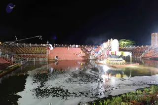 Night photograph of a illuminated temple tank at an ashram. A large lotus-shaped pavilion decorated with string lights, yellow and red-green cloth drapes and a kalasha-like top stands on a raised platform in the water at right. Terraced red-brick steps lead down to the water on the left and centre, with a few small figures in white and light clothing standing along the steps and railings. Strings of lights line the terraces, and a tall lit tower stands at the far right edge. Water is dark and reflective, with floating vegetation. Several figures are visible but small and partly in shadow.