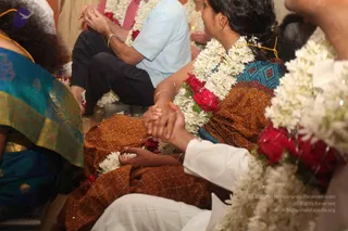Close photo of seated devotees at a ceremony, most of them wearing white jasmine garlands and red rose garlands. In the centre, a woman in a brown embroidered top and a blue patterned blouse holds hands with a man in white clothing, their clasped hands resting on her lap. To the left, a woman in a teal and gold sari sits beside a man in a pale blue shirt and dark trousers. Gold bangles and a gold necklace are visible. The scene is crowded and only partly visible at the frame edges.