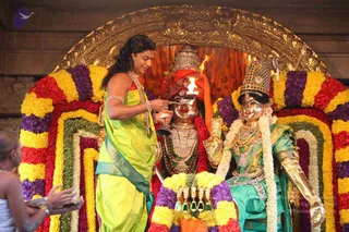 A photograph of a temple ritual before a deity shrine framed by arches of marigold and rose garlands in yellow, purple, red and green. A person with long dark hair and a red tilak, wearing a lime-green silk sari with a teal border and a pearl-and-gold necklace and bangles, holds a brass lamp with a bright flame and a bell-shaped vessel before two metal deity murtis. The central murti wears a crown and a red cloth, and the right murti wears an ornate jeweled crown and a green silk garment with gold armlets. A bare-chested assistant in white dhoti stands at the lower left, holding a plate.