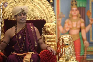 A seated man sits on an ornate gilded throne with a deep red cushion, wearing a cream turban with a jewelled ornament, a plum-coloured dhoti-style garment with gold border and a shawl draped over the shoulder, a long rudraksha mala, and bronze-coloured bangles. He smiles and rests one hand on a red velvet armrest beside a large gold lion statue. Behind him, a painted deity murti with multiple arms and a crown stands in a brightly coloured temple setting with orange and green tones.