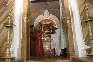 A photograph of a temple shrine doorway with polished brown-and-cream marble walls and two tall brass oil lamps flanking the entrance. Inside, a glowing orange arch frames a small deity murti decorated with flowers on a stone pedestal. Three men stand at the shrine: one in a red-orange dhoti with a shawl-like cloth and a bare upper body, bowing with hands near the murti; another in a white dhoti and shawl, bare-headed and bare-chested, leaning in with hands extended toward the deity; and a third in a red-orange garment with a garland at the left edge. A green curtain hangs to the right of the doorway.