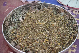 A close-up photograph of a large round stainless steel plate filled with a heaped mixture of dried herbs, bark pieces, twigs, seed husks, dried flower petals and small grey stones or nuts. The plate rests on a red-brown floor beside a purple and yellow patterned cloth with a fringed edge, and a white and red fan-like item is visible at the top right. No people are visible. The mixture appears to be prepared botanical material for a ritual offering.
