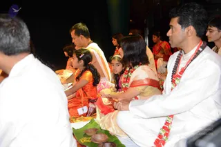 Devotees seated in rows during a pada-puja style ritual indoors at night. In the foreground a man in white kurta with a red and white garland and rudraksha-style beads sits beside a woman in a cream and gold sari with a red border and red garland, holding a small offering. Beside them a girl in an orange dress and a younger girl in pink and a white flower in her hair watch attentively. A banana leaf with small clay oil lamps lies at the bottom. A man in a white shirt sits on the left edge, and a woman in red and gold sits behind, with others in the background.