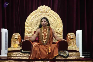 A man with long swept-back dark hair, a red tilak on his forehead, a headset microphone, and an orange-saffron robe with a gold-coloured wrap sits on an ornate gold throne with a tall arched back and two gold lion statues at its base. Heavy gold beaded necklaces and a rudraksha-style mala hang on his chest, with gold armbands on his upper arms. Dark maroon velvet drapes form the backdrop, with white tower fans on either side. The appearance of the seated man with long hair, saffron robe, and ceremonial gold ornaments is a probable Swamiji match, and he is shown on a dais in a satsang-style hall.