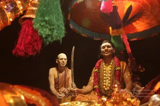Photo of a procession at night beneath an ornate umbrella canopy with red, green, orange and purple tassels. A man with long dark hair, a gold necklace, a red and pink flower garland and orange robes sits on a golden seat, holding a ceremonial sword upright, with a golden bowl in the foreground. Beside him sits a shaved-headed man in saffron robes and a rudraksha mala, holding a small object in his hands. Gold ornaments and lamps glow around them. Swamiji (probable) is the central figure seated with the sword, shown with long hair, a gold necklace, a garland and saffron robes.