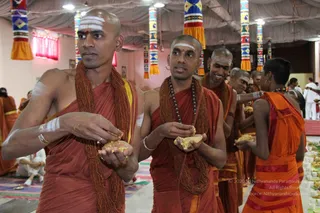 A group of shaved-headed men in saffron-orange robes stand in a line inside a decorated hall with patterned cloth hangings and tasselled pillar covers. Several hold food offerings in their cupped hands, and those in front wear rudraksha malas and white urdhva pundra tilak on their foreheads. Behind them, others in orange dress stand near tables of prepared offerings, and in the background a person in white clothing stands near a raised platform. The scene appears to be a ritual food offering or prasadam distribution. No person is clearly identifiable as a person from the visible appearance.