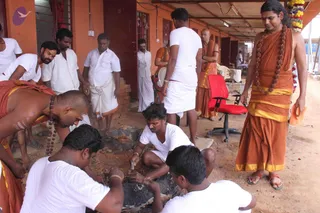 A photograph of an outdoor temple-work scene beside an orange-painted building with a covered walkway. Several men in plain white shirts and white dhotis crouch and kneel on bare earth around a dark stone block, working on it with chisels and tools; one bare-chested man in orange cloth with a rudraksha mala leans over the stone on the left. Behind them, a standing man in orange robes with a rudraksha mala and a shaved head is visible near a red office chair. On the right, a tall man with long swept-back hair wears an orange dhoti, a yellow-bordered orange shawl, a long rudraksha mala and orange sandals, holding an orange cloth, and appears to be watching the work; a flower garland hangs at the top right. Appearance-based probable candidate: Swamiji (probable), standing at right with long hair, rudraksha mala and orange robes, observing the stone carving.