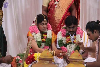 A South Indian wedding ritual photograph. A bride in an orange and gold silk sari with gold jewellery and a maang tikka sits with her palms joined over a brass vessel, next to a groom in a white dhoti with a gold-bordered shawl. Both wear pink, white and green flower garlands. Behind them stands a woman in a red and gold saree, and at right a priest with a shaved forehead marking and bare shoulders performs the ceremony. Brass kalasha pots, flowers and a bundle of grass lie in the foreground against a white draped backdrop.