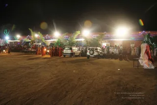 Night photograph of an outdoor ashram courtyard of packed dirt, lit by bright floodlights and strung with multicoloured fairy lights along a low terracotta-roofed building. Several saffron-robed figures in orange and red garments walk in a loose group across the centre, some carrying small lit lamps. Two white SUVs are parked in the middle distance. A crowd of people in white and light clothing stands along the building's front and right side watching. Potted green trees are decorated with lights, and a folded white and orange cloth lies on a table at the far right edge.