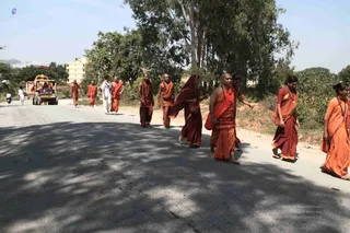A photograph of a procession walking along a sunlit paved road lined with trees and a yellow building. Roughly a dozen people in saffron and orange robes walk in a line, many barefoot. A man in the centre-right wears an orange shawl over bare shoulders and a red-orange dhoti, with a beaded mala around his neck. Behind him, a woman in a deep maroon sari with a red shawl walks beside others in orange. On the right, two women in orange saris wear rudraksha malas. A small orange-and-gold decorated cart or vehicle with a canopy is visible at the left edge, with a man in white standing near it. Shadows of trees fall across the road.