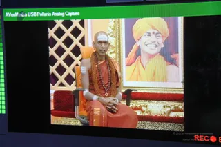 A screen capture from an AVerMedia USB Polaris Analog Capture window shows a live video feed. A bald man with white vertical tilaka marks and wrapped white arm bands sits on an ornate orange chair, wearing orange robes, a rudraksha mala and a gold-beaded necklace, hands folded in his lap. Behind him hangs a gilded framed portrait of a smiling man in a yellow-orange turban and yellow-orange robes. The chair and bench are cushioned in red with gold trim, and a lattice screen stands at the left. The capture overlay shows a green bar and a small red recording indicator in the corner.