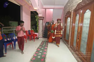 A photograph taken at night inside a pink-walled house in Tamil Nadu. Three men in saffron and orange-red robes walk along a tiled corridor toward the camera. The man at right wears a red-orange turban and a long garland of white and red flowers, and gestures with raised hands in greeting. Another man beside him wears a saffron robe and a rudraksha-style beaded necklace. A young boy in a pink shirt and red trousers on the left stands with palms pressed together in namaskar. A path of flower petals and rose-coloured blossoms lines the floor ahead, and blue, red and white plastic chairs sit along the left wall. Carved wooden doors with frosted glass panels stand on the right.