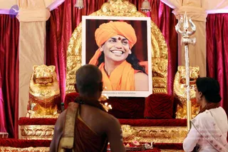 A photograph of a ceremonial dais draped in red and cream fabric with a golden throne-like seat flanked by two golden lion figures. At the centre a large printed portrait shows a smiling man in an orange turban and orange robe with a red bindi and long dark hair, its caption reading 'Paramahamsa Nithyananda'. A silver trident stands at the right edge. In the foreground a shaved-headed devotee in a rust-coloured robe with a beaded necklace faces the portrait, and a woman in a white sari with a dark plait sits to the right. Offerings and a small pink flower rest on the dais.