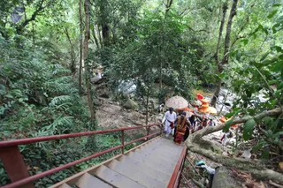 A photograph taken from above a red-railed concrete staircase descending through dense green forest toward a rocky stream. At the bottom of the steps, a small group walks along the rocky bank, including a figure in white dhoti and shirt and a person in red and white garments wearing a red-and-orange garland, with other people in dark and light clothing behind. Beside them a large round decorated umbrella or canopy and yellow and orange items are visible near the water. Another figure in white stands further along the path among the trees and rocks.