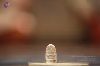 A small clear crystal lingam stands on a light flat pedestal in the foreground, its surface carved with horizontal bands and geometric triangular patterns. The background is an out-of-focus warm blend of brown, cream and reddish tones, with no people visible.