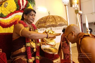 A photograph of a ceremonial scene with a seated figure in dark maroon and brown robes, wearing long red and white flower garlands, a gold necklace and gold armlets, extending one hand over a silver plate. In front, a bald man in saffron-orange robes with a rudraksha mala bends forward, wearing glasses. Behind the seated figure stands a large gold ceremonial umbrella with fringe and a gold kalasha, flanked by tall silver staffs. A floral arrangement of yellow, orange, red and white flowers decorates the background, with a second attendant in maroon at right.