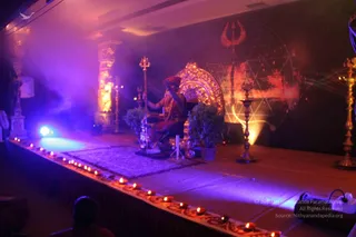 A stage lit in violet and warm amber for an evening event. A seated figure in orange-red robes and a red turban sits on an ornate gold-backed throne, raising one hand, with a tall brass trident-topped staff beside them. A row of small oil lamps lines the front edge of the stage, and brass lamp stands and potted plants flank the throne. The backdrop shows a red trident and a glowing sri yantra graphic with a smoky haze in the air. A dark silhouette of a seated audience member sits at the lower left. The seated figure is a distant, partly backlit figure whose face is not clearly visible, so identity remains uncertain.