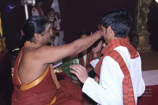 Indoor ritual scene in a maroon-draped hall. A person in a rust-red saffron-toned robe with a gold-edged wrap and a gold beaded necklace sits on the left, raising an arm and touching the forehead of a young man in a white kurta with an orange scarf printed with script. Behind them, a brass oil lamp and an ornate gold pillar stand near other seated attendees in white and red. The seated person has a red flower in their hair and a tilak on the forehead, and the young man presses his palms together in respect.