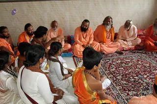 A seated group in a plain room with a red patterned rug and a light woven wall. Several saffron-robed sadhus and men with long grey or white beards and ash markings sit on the floor, some with rudraksha malas. On the left, young students in white with gold-bordered orange cloth and beaded necklaces sit with their backs partly to the camera, some holding papers. A man in a dark-bearded saffron robe sits centre-right, and an older bearded sadhu in pale orange gestures in conversation. The mood is an attentive gathering with one speaker addressing the circle.