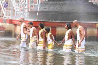 A photograph of six young men walking in a line through shallow water in a pool beside a large dark stone structure with carved lotus-petal patterns. They wear white dhotis bordered with gold, bare-chested, and hold hands as they wade. The bald man at the left wears glasses. Water spray arcs from a fountain at the top, and a metal staircase and red-and-white painted pool wall are visible behind them.