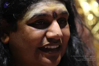 Close-up photograph of a smiling person with long dark wavy hair, a yellow-ochre painted forehead and a dark red bindi-style mark between the brows, showing a nose stud. Blurred golden bokeh lights fill the background on the right. The face fills most of the frame and no other people are visible.