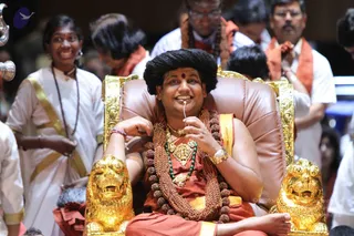A photograph of a man seated on a brown leather throne with gold lion-head armrests, wearing a saffron-orange and gold dhoti-style cloth, multiple rudraksha mala necklaces, a green bead strand, a gold pendant and a gold wristwatch, with a red bindi on his forehead and long dark hair. He smiles while holding a small white object near his mouth. Around him, a woman in white with a gold-bordered cloth and beaded mala stands at left, and several men wearing white shirts with saffron-orange shawls stand behind him. The setting appears to be a hall with a partial golden railing or metal fixture at the upper left.