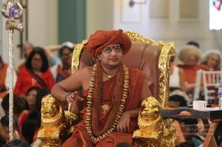A seated man in saffron-orange robes and an orange turban sits on an ornate gold-and-brown throne with carved golden lion-head armrests. He wears a long gold-bead garland over rudraksha malas and a gold pendant necklace, and holds one hand raised near the armrest while looking upward. Behind him, a seated audience in orange and red clothing fills the hall. A white cup rests on a small side table with an orange-trimmed tray, and a silver ceremonial staff stands at the left edge. Swamiji (probable), seated on the throne in saffron robes, turban and rudraksha malas, presenting a discourse before the audience.