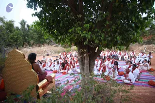 Outdoor photo of a seated audience under a large leafy tree on a colourful striped cloth. About forty people in white clothes with orange-red scarves sit in rows facing a gilded throne-like seat at the left, where a figure with long dark hair, in an orange-red garment, sits with back partly to the camera. A microphone stands near the throne. Some attendees hold orange cloth bundles, and dry scrubby ground and trees surround the gathering.