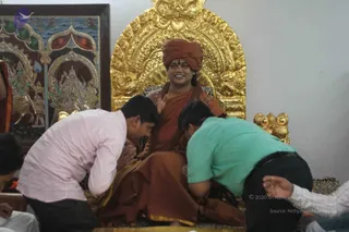 A seated figure in an orange-red turban and saffron-orange robe sits on an ornate gold throne with carved lion-like finials, smiling broadly, with a red tilak on the forehead and a long mala around the neck. Two men in front bend forward with hands pressed together, one in a pink shirt and one in a teal shirt and dark trousers, receiving or offering blessings. Behind the throne hangs a framed painting of Hindu deities with mountains and figures in gold-and-red tones. The lower right shows another person's hand near the floor. Appearance-based label: a person, with a turban and orange robes and rudraksha-style beads, seated on the throne as the one being honoured.
