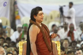 A man with long dark hair swept back stands beside a gold-trimmed brown leather throne in front of a large seated crowd under a white tent. He wears a rust-orange saffron wrap over his shoulder and a thick rudraksha mala draped around his neck, with a white ash mark on his forehead, and gestures with his right hand while speaking. Behind him a cameraman in white stands near a tripod, and many devotees in white and orange clothing sit in rows.