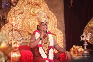 A photograph of a seated man on an ornate gold throne with elaborate repoussé carvings, wearing a tall gold crown with a green stone, a red-brown robe, layered gold necklaces and gold bangles. He wears a white and pink flower garland around his neck, has a red bindi on his forehead, and raises one hand in a blessing gesture while smiling. A golden trident (trishul) stands at left, a golden lion-head ornament sits near his feet on the right, and a silver peacock-shaped stand appears at the far right beside red velvet cushions and a dark red curtain. The setting suggests a ceremonial throne presentation.