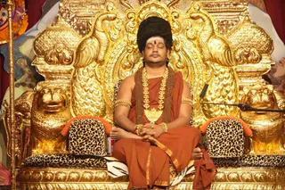 A seated man with eyes closed sits cross-legged on an ornate golden throne carved with peacocks and lion heads, upholstered with leopard-print cushions and a tiger-striped cloth. He wears a saffron-orange robe with gold border, a heavy gold necklace with a gold pendant, rudraksha beads, gold armlets and bangles, and a sweeping upswept dark hair style with a red tilak on his forehead. His hands are pressed together in his lap. A microphone stand and a ceremonial staff stand beside the throne, with colourful painted backdrop behind. a person is seated as the presiding figure, identified by his long swept hair, saffron robe, ornate gold necklaces and rudraksha malas.