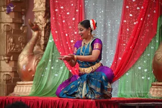 A female classical dancer kneels on a red-draped stage, smiling and extending her right hand palm-up in a gesture. She wears a teal and purple silk sari with gold zari borders, a gold waist belt, gold bangles, a necklace, earrings, a nose ring and a white and red flower garland in her hair. Behind her hang red and green draped fabrics with white dot patterns, and on the left stands an ornate golden carved pillar with a sculpted swan-like figure. No a person candidate is visible.
